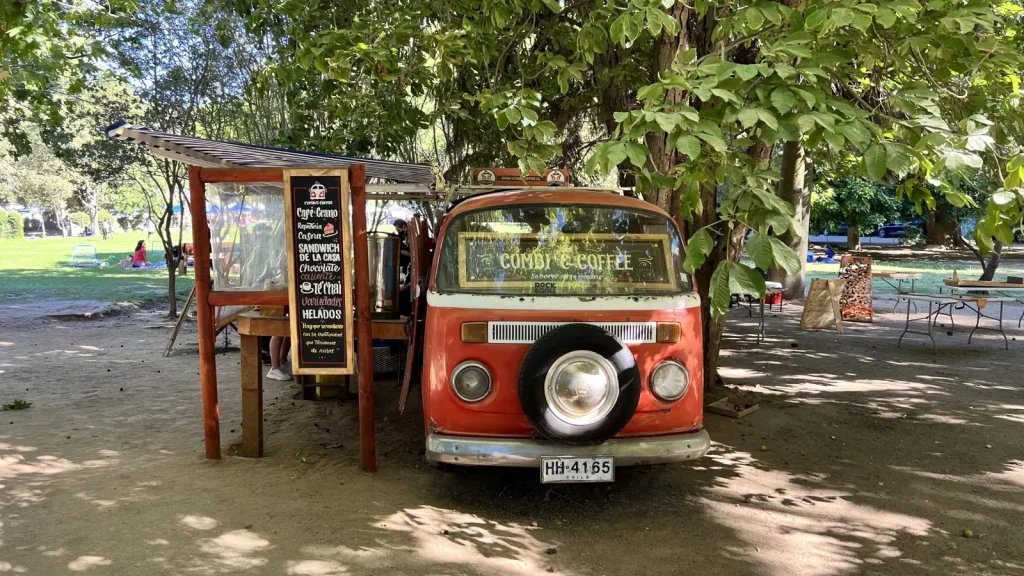Furgón rojo de Combi Coffee bajo los árboles, con su toldo y su menú