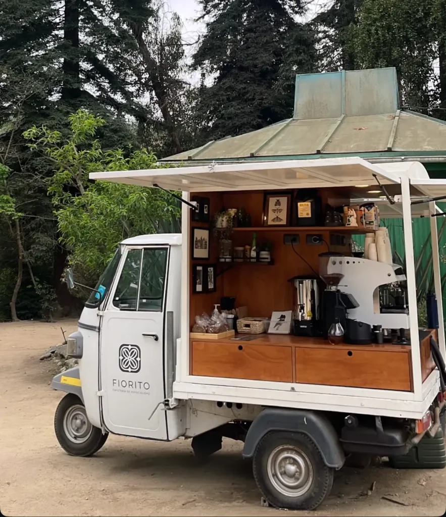 Carrito blanco de Fiorito Cafetería de Especialidad con la vitrina abierta, máquina de espresso y repisas de madera, bajo los árboles